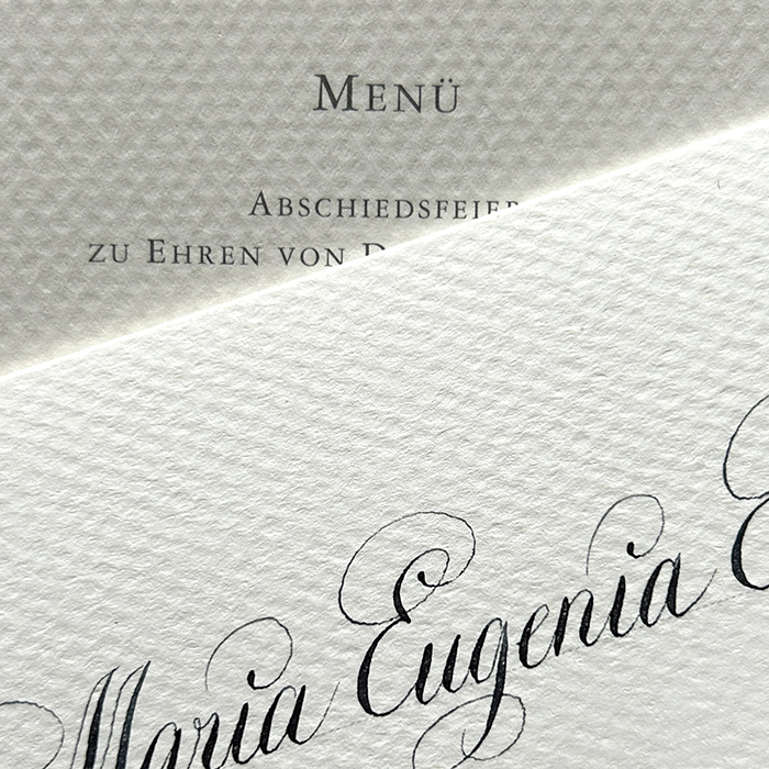 Detailaufnahme einer hochwertigen Einladungssituation mit kalligrafisch geschriebenem Namen auf edlem Papier, im Hintergrund eine angeschnittene Menükarte